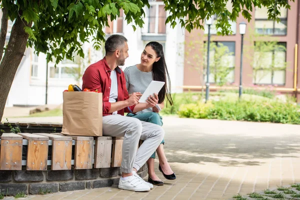 Seitenansicht von Eheleuten, die gemeinsam ein Tablet benutzen, während sie sich nach dem Einkaufen auf einer Bank auf der Straße ausruhen — Stockfoto