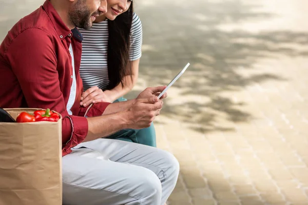 Partial view of married couple using tablet together while resting after shopping on bench on street — Stock Photo