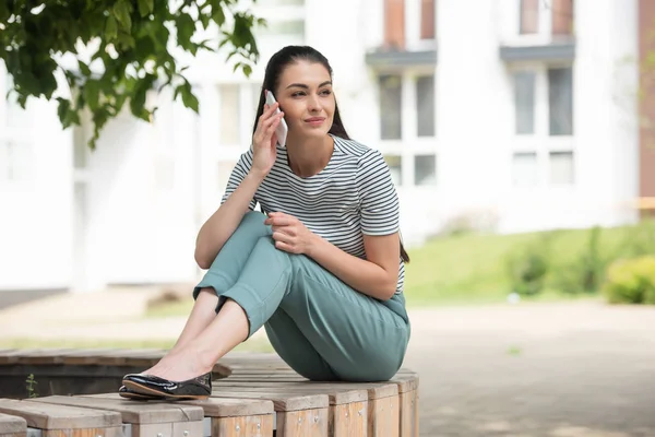 Красивая женщина разговаривает по смартфону, отдыхая на деревянной скамейке на улице — стоковое фото