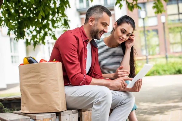 Vista laterale della coppia sposata utilizzando tablet insieme durante il riposo dopo lo shopping sulla panchina sulla strada — Foto stock