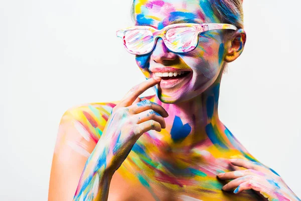 Chica atractiva sonriente con colorido arte corporal brillante y gafas de sol tocando los labios aislados en blanco - foto de stock