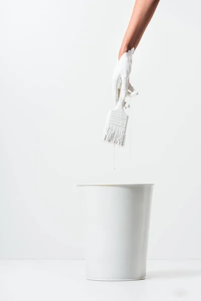 Imagen recortada de la mujer sosteniendo pincel de pintura con pintura blanca por encima de cubo en blanco - foto de stock