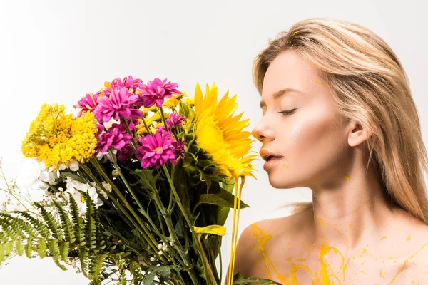 Vista lateral de mujer atractiva con pintura amarilla en el cuerpo olfateando flores aisladas en blanco - foto de stock