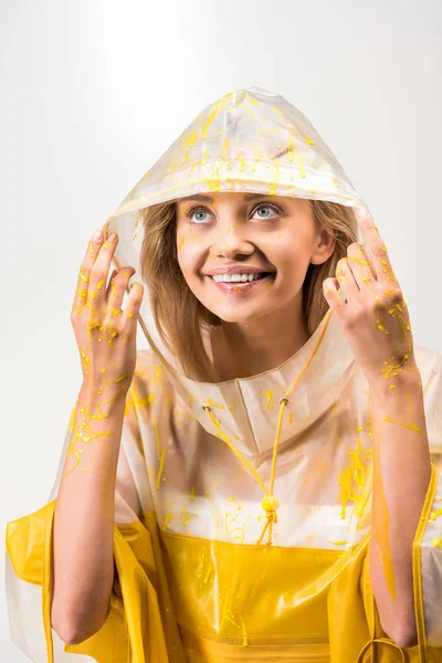 Mujer sonriente en impermeable pintado con pintura amarilla tocando la capucha y mirando hacia arriba aislado en blanco - foto de stock