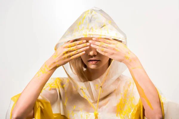 Girl in raincoat painted with yellow paint covering eyes with hands isolated on white — Stock Photo