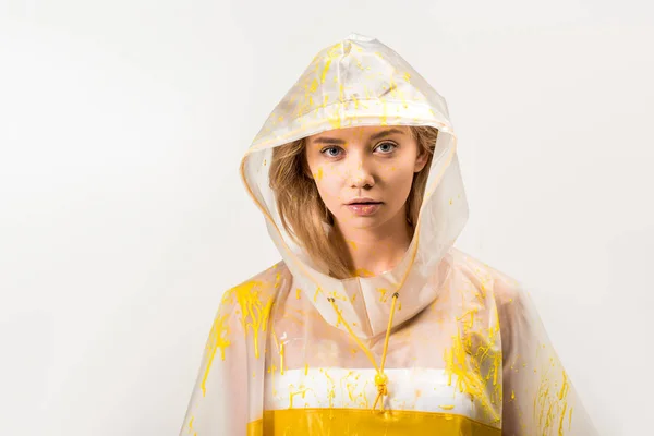 Attractive woman in raincoat painted with yellow paint looking at camera isolated on white — Stock Photo