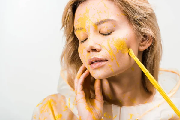 Headshot of beautiful woman in raincoat painting face with yellow paint isolated on white — Stock Photo