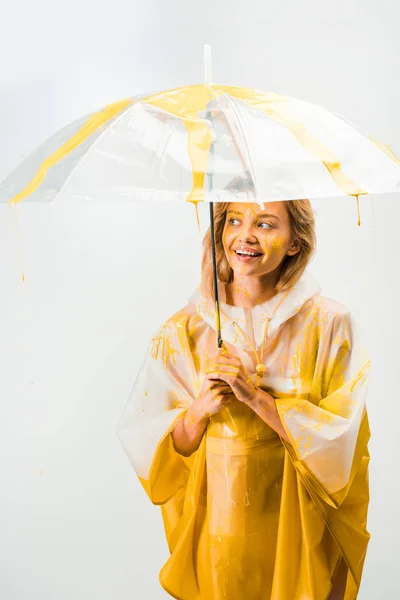 Mujer feliz en impermeable pintado con pintura amarilla de pie bajo paraguas aislado en blanco - foto de stock