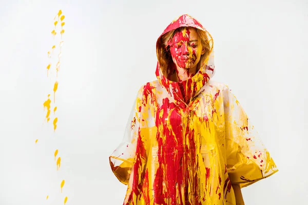 Atractiva mujer en impermeable pintado con pinturas amarillas y rojas de pie con los ojos cerrados aislados en blanco - foto de stock