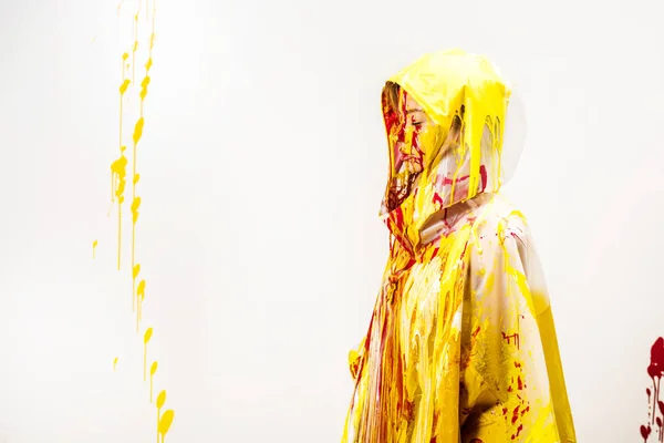 Vista laterale della donna in impermeabile verniciato con vernici gialle e rosse in piedi in cappuccio isolato su bianco — Foto stock