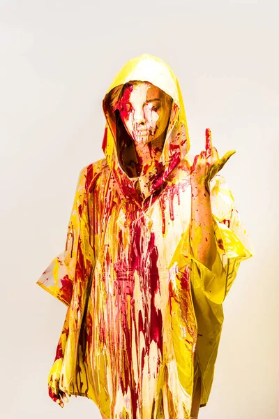 Schöne Frau im Regenmantel, bemalt mit gelben und roten Farben, die den Mittelfinger isoliert auf weiß zeigen — Stockfoto