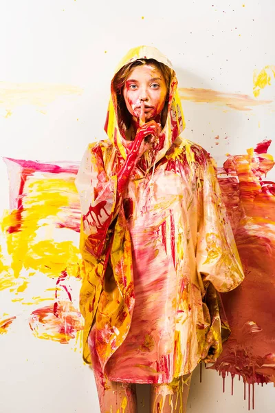 Beautiful woman in raincoat painted with yellow and red paints showing silence gesture — Stock Photo