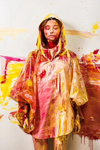 Bela mulher em capa de chuva pintada com tintas amarelas e vermelhas de pé com olhos fechados — Fotografia de Stock