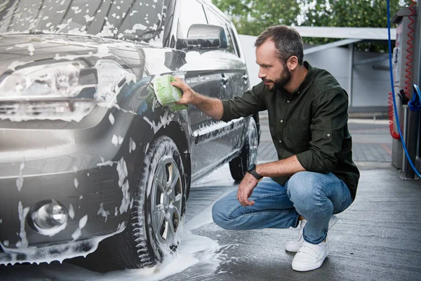 Schöner Mann hockt und putzt Auto in Waschanlage mit Lappen und Schaum — Stockfoto