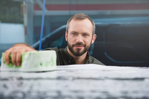 Schöner lächelnder Mann reinigt Auto in Waschanlage mit Lappen und Schaum — Stockfoto