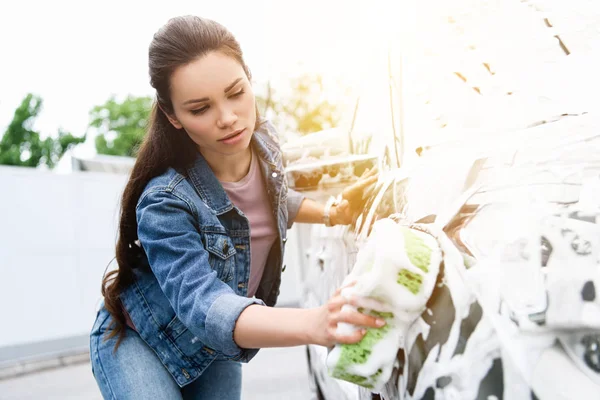 Schöne Frau putzt Auto in Waschanlage mit Lappen und Schaum — Stockfoto