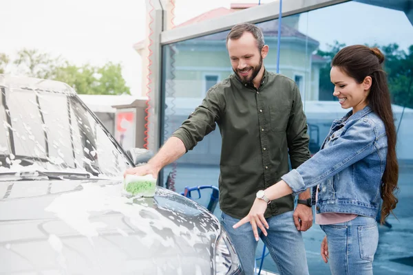 Novia apuntando en coche a novio mientras él la limpieza en el lavado de coches - foto de stock