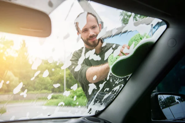 Hombre guapo limpiando la ventana delantera del coche con trapo y jabón en el lavado de coches - foto de stock