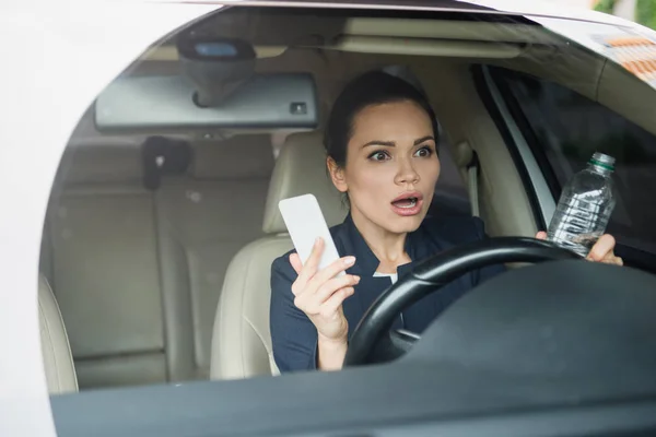 Chocado atraente mulher segurando smartphone e garrafa de água durante a condução do carro — Fotografia de Stock