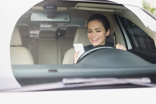 Mujer atractiva sonriente usando teléfono inteligente mientras conduce el coche - foto de stock