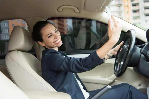 Attractive smiling driver taking selfie with smartphone in car — Stock Photo