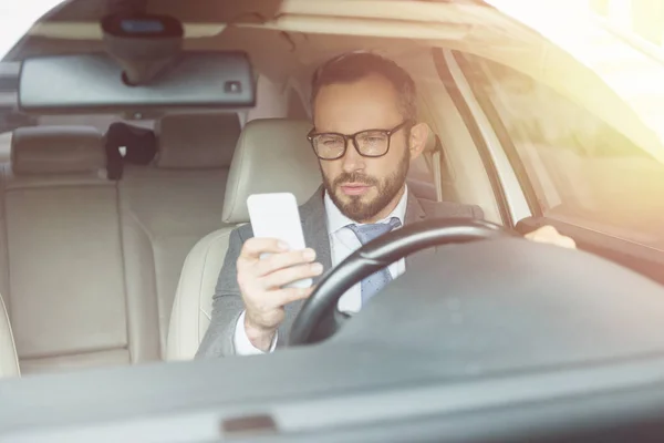 Hombre de negocios guapo usando el teléfono inteligente mientras conduce el coche - foto de stock