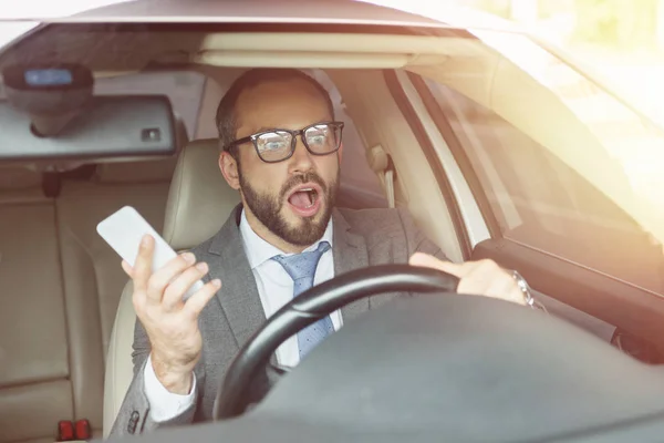 Schockierter schöner Mann hält Smartphone in der Hand und schaut im Auto weg — Stockfoto