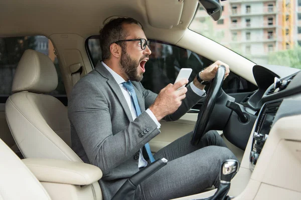 Side view of handsome businessman screaming in car and holding smartphone in hand — Stock Photo