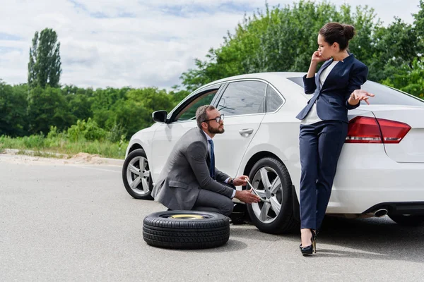 Handsome businessman changing tires on car on road, businesswoman talking by smartphone — Stock Photo