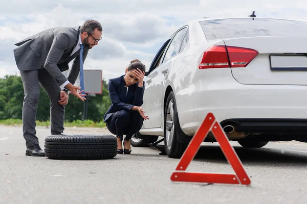 Businessman gesturing and sad businesswoman squatting near broken car on road — Stock Photo