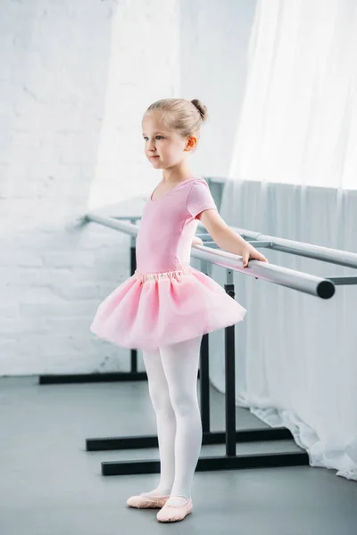 Vista lateral del niño en tutú rosa practicando ballet y mirando hacia otro lado en el estudio de ballet - foto de stock