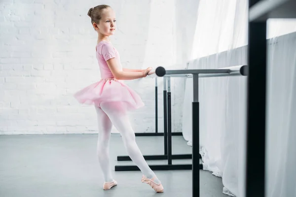 Vista lateral de la adorable bailarina en tutú rosa practicando ballet y mirando hacia otro lado - foto de stock