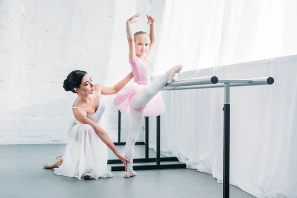 Adorable niño en tutú rosa estiramiento y mirando a la cámara mientras practica ballet con el maestro - foto de stock