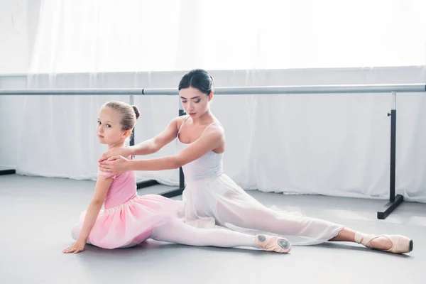 Adorable little ballerina in pink tutu looking at camera while exercising with teacher in ballet school — Stock Photo