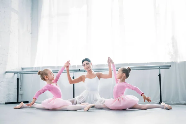 Joven profesor de ballet con niños en faldas tutú sentados y tomados de la mano mientras hace ejercicio en la escuela de ballet - foto de stock