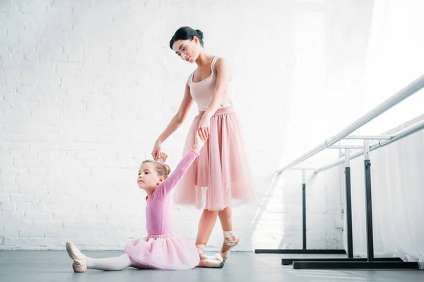 Молодая учительница балета смотрит на ребенка в розовой пачке, растянувшегося в балетной школе — стоковое фото
