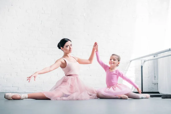 Hermosas bailarinas adultas sonrientes y pequeñas bailarinas estirándose y tomándose de la mano en el estudio de ballet - foto de stock