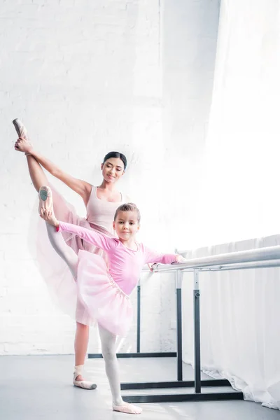 Sorrindo jovem professor de balé olhando para pouco estudante alongamento e olhando para a câmera no estúdio de balé — Fotografia de Stock