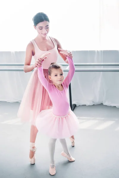 Entzückendes Kind im rosafarbenen Tutu blickt in die Kamera, während es mit einem Lehrer in der Ballettschule trainiert — Stockfoto