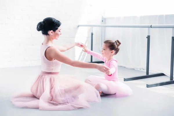 Vista lateral del profesor de ballet y pequeño estudiante en faldas de tutú rosa sentado y cogido de la mano mientras se entrena en la escuela de ballet - foto de stock