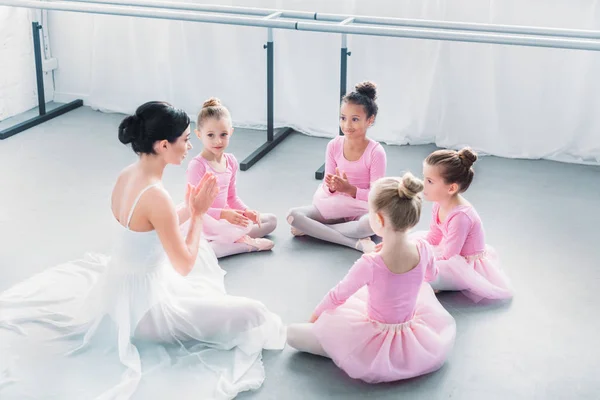 Высокий угол обзора детей в розовых юбочках и молодой учитель балета, сидящих вместе в балетной школе — стоковое фото
