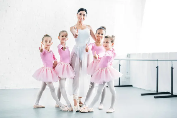 Pequeñas bailarinas y profesora de ballet mostrando los pulgares hacia arriba y sonriendo a la cámara en la escuela de ballet - foto de stock