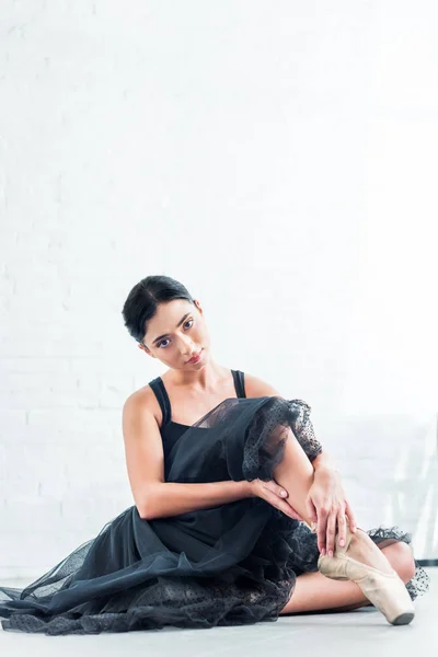 Hermosa joven bailarina en tutú negro sentado y mirando a la cámara en el estudio de ballet - foto de stock