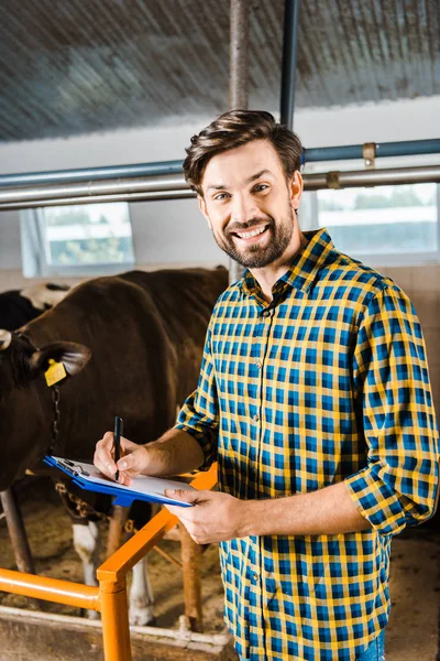 Fazendeiro bonito verificando vacas em prancheta estável e segurando com documentos — Fotografia de Stock