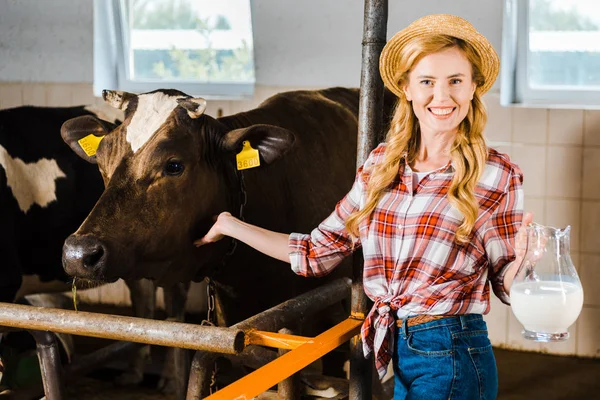 Attractive farmer holding jug of milk and palming cow in stable — Stock Photo