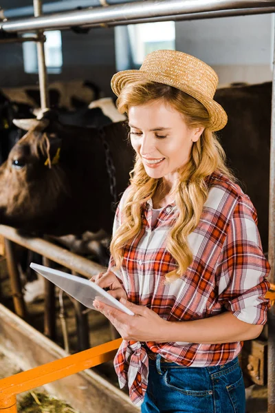 Atractivo granjero sonriente en sombrero de paja usando tableta en establo - foto de stock