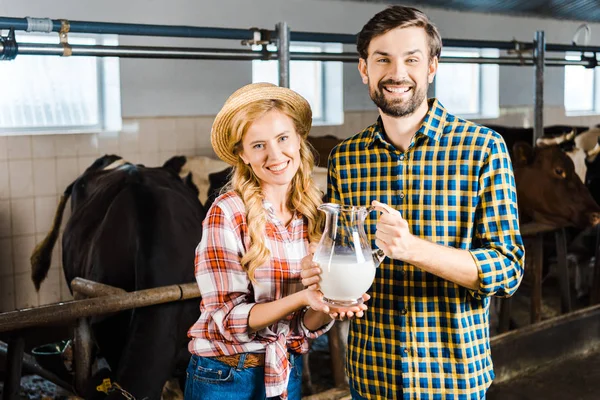 Feliz pareja de agricultores sosteniendo jarra de leche en establo - foto de stock