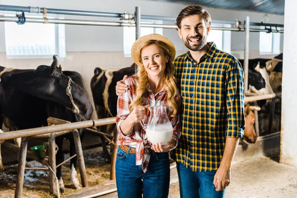 Casal sorridente de agricultores segurando jarro de leite em estábulo com vacas — Fotografia de Stock