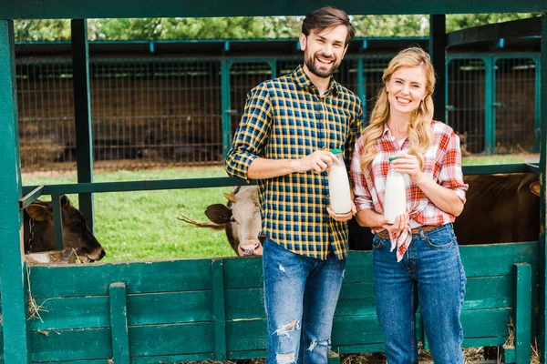Sonrientes parejas de granjeros sosteniendo botellas de leche de vaca cerca de establo - foto de stock