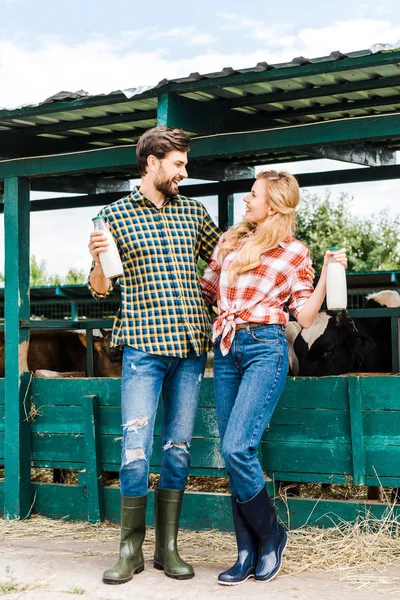 Couple of farmers holding bottles of cow milk near stable and looking at each other — Stock Photo